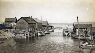 Original Mackinaw boats are moored on the south (left) side the river. The first generation of enclosed fish tugs are tied up on the north side.